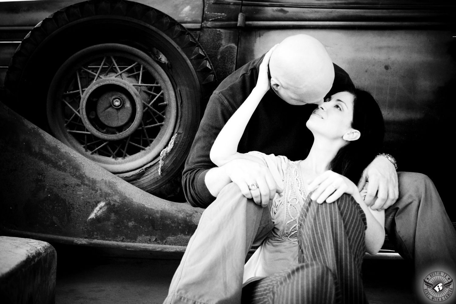 Dark haired Italian lady in lacy white flowing blouse and grey and black striped dress pants gazes in the eyes of a guy with a shaved head wearing a black long sleeve turtle neck shirt sit on the  running board of a vintage  car next to a ripped spare tire in this nostalgic engagement photo in Taylor Texas.